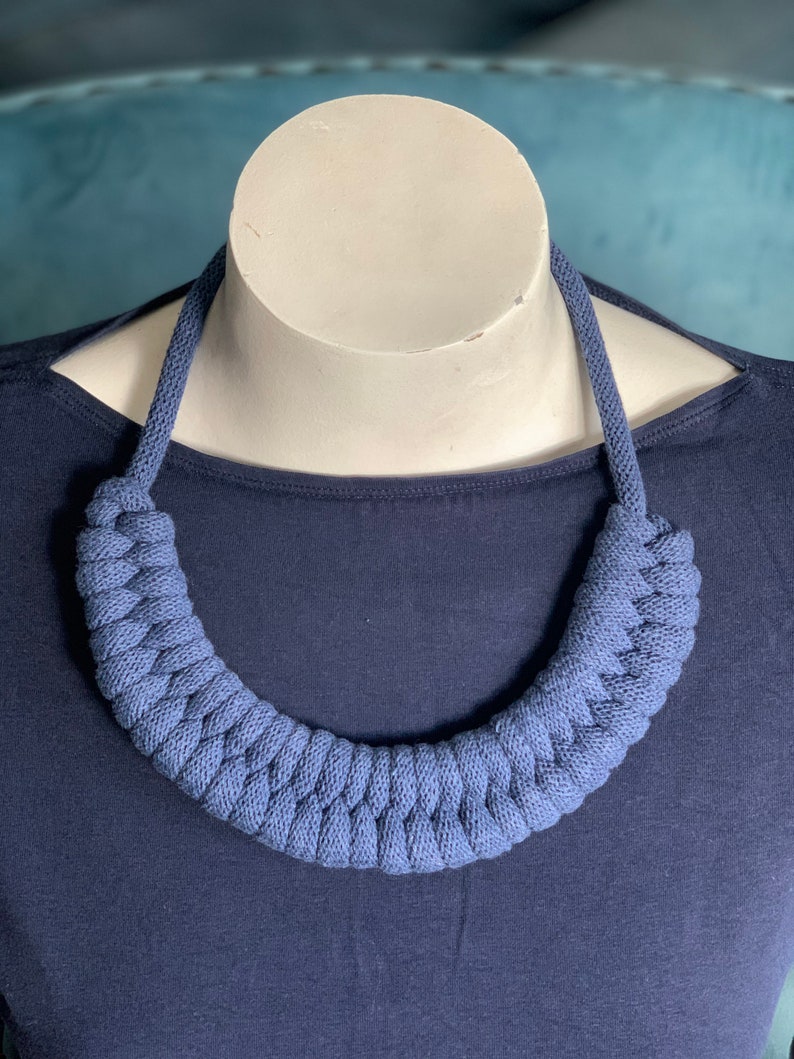 Op de afbeelding: Een blauwe gebreide ketting die wordt getoond op een witte mannequinhoofd. De ketting heeft een dik, gevlochten ontwerp in een effen blauwe kleur. De mannequin staat tegen een donkerblauwe top.