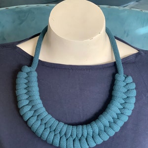 Op de afbeelding: Een turquoise gevlochten ketting, gepresenteerd op een witte mannequinhoofd. De ketting is gemaakt van een dik, geweven materiaal, waardoor hij een dikke, getextureerde uitstraling heeft. De achtergrond is effen marineblauw, wat contrasteert met de ketting.
