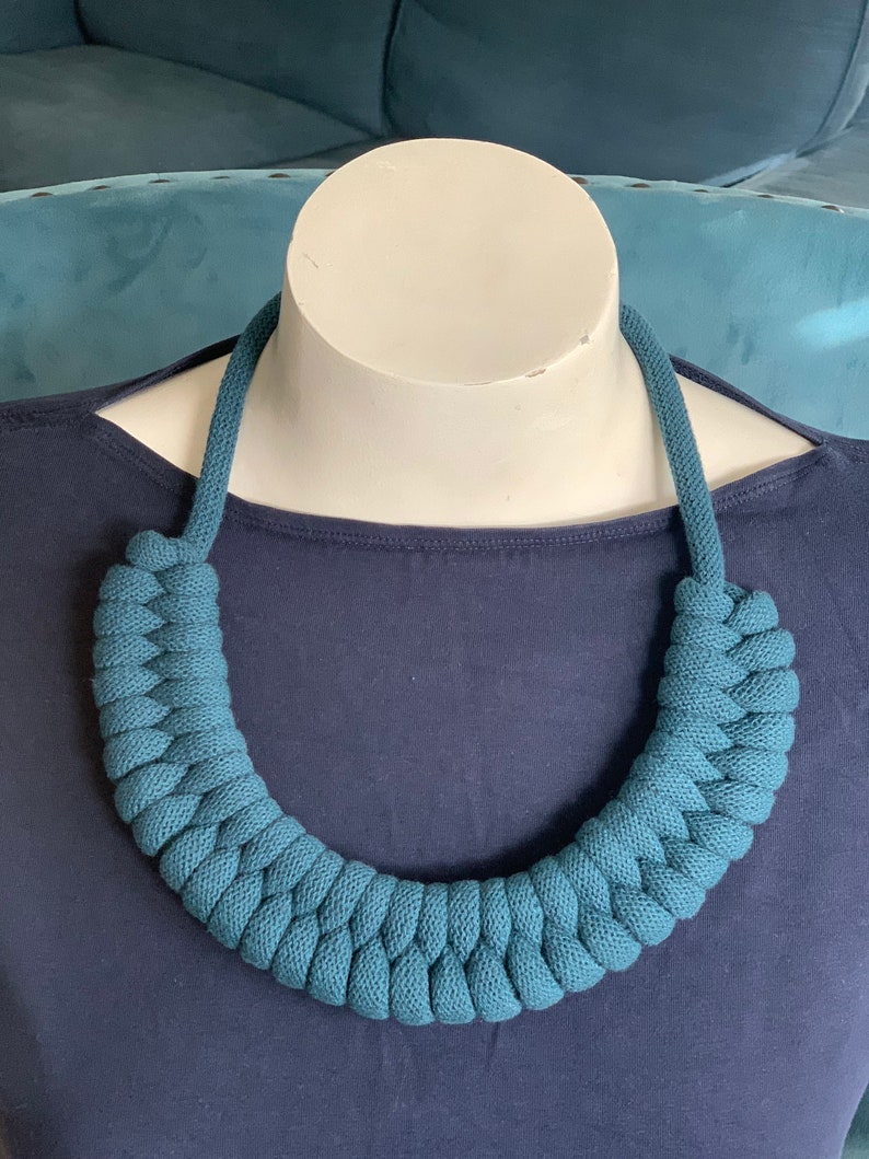 Op de afbeelding: Een turquoise gevlochten ketting, gepresenteerd op een witte mannequinhoofd. De ketting is gemaakt van een dik, geweven materiaal, waardoor hij een dikke, getextureerde uitstraling heeft. De achtergrond is effen marineblauw, wat contrasteert met de ketting.