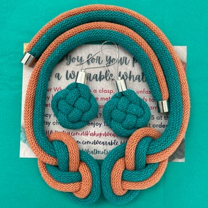 May include: A teal and peach-colored knotted necklace and matching earrings set. The necklace and earrings are made of a thick, woven material. The earrings have silver-colored metal accents. The background is a solid teal color.