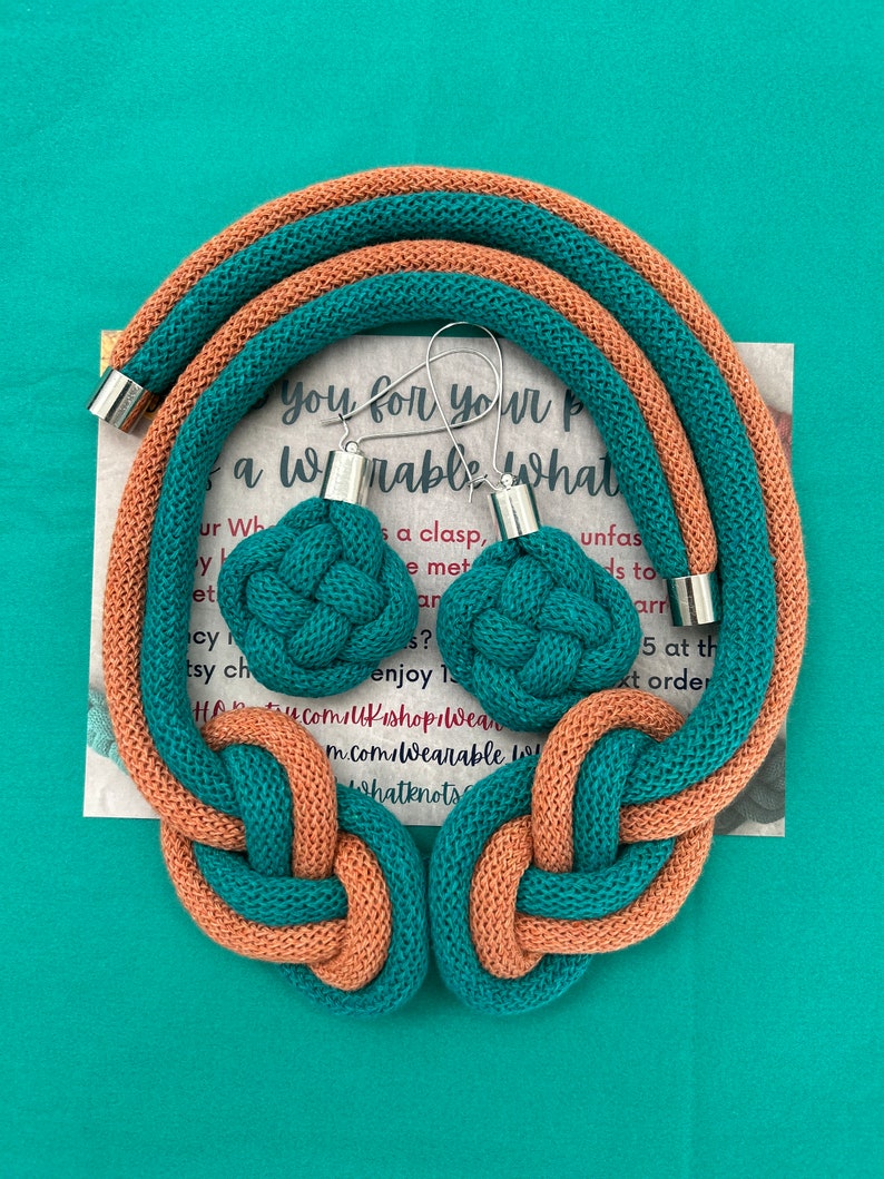 May include: A teal and peach-colored knotted necklace and matching earrings set. The necklace and earrings are made of a thick, woven material. The earrings have silver-colored metal accents. The background is a solid teal color.