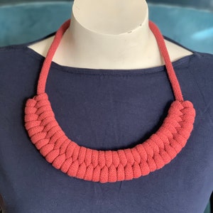 Op de afbeelding: Een terracotta-kleurige, gevlochten gebreide ketting. De ketting wordt getoond op een witte mannequinhoofd tegen een donkerblauwe achtergrond. De ketting heeft een dik, afgerond ontwerp en een geweven textuur.