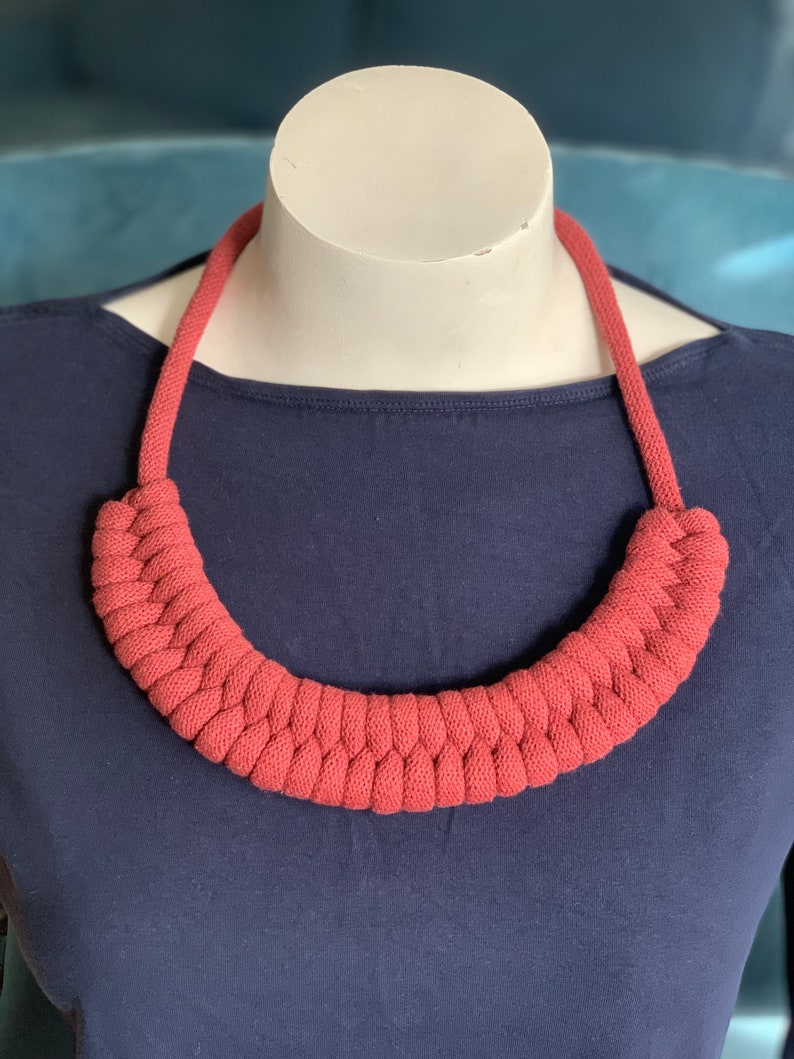 Op de afbeelding: Een terracotta-kleurige, gevlochten gebreide ketting. De ketting wordt getoond op een witte mannequinhoofd tegen een donkerblauwe achtergrond. De ketting heeft een dik, afgerond ontwerp en een geweven textuur.