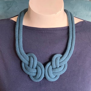 May include: Teal-colored knotted rope necklace. The necklace features two large, symmetrical knots at the front, connected by a double strand of rope that goes around the neck. The necklace is displayed on a mannequin.