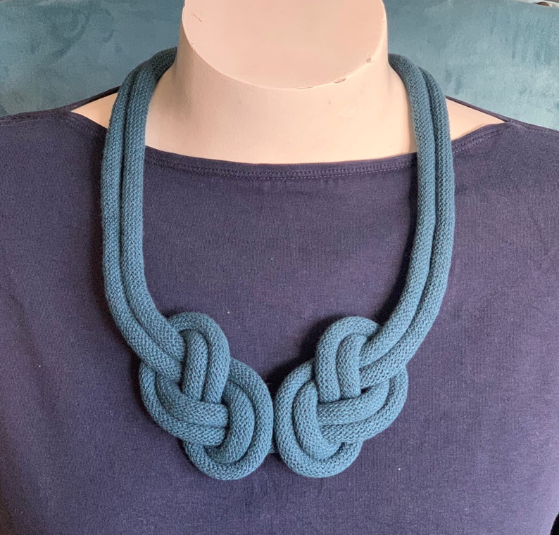 May include: Teal-colored knotted rope necklace. The necklace features two large, symmetrical knots at the front, connected by a double strand of rope that goes around the neck. The necklace is displayed on a mannequin.