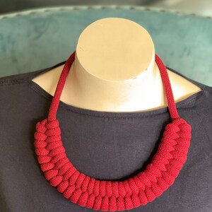 Op de afbeelding: Een opvallende, rode gebreide ketting op een mannequin. De ketting heeft een dik, gevlochten ontwerp rond de nek, met een eenvoudig koord voor de rest van de ketting. De achtergrond is een donkerblauwe top en een lichtblauwe ondergrond.