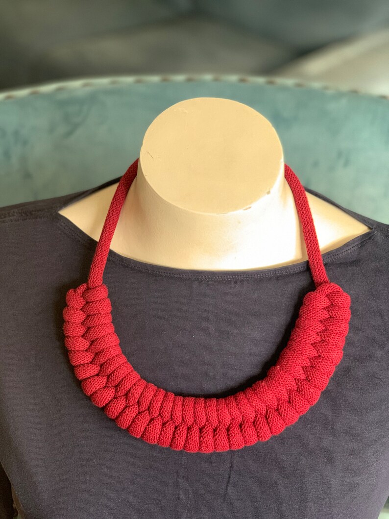Op de afbeelding: Een opvallende, rode gebreide ketting op een mannequin. De ketting heeft een dik, gevlochten ontwerp rond de nek, met een eenvoudig koord voor de rest van de ketting. De achtergrond is een donkerblauwe top en een lichtblauwe ondergrond.