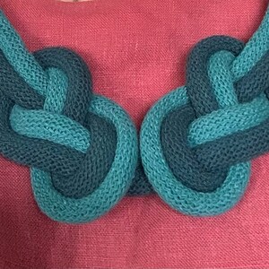 May include: A close-up of a knotted necklace in shades of teal and navy blue. The necklace is made of a thick, textured material and rests against a pink background. The necklace is a statement piece.