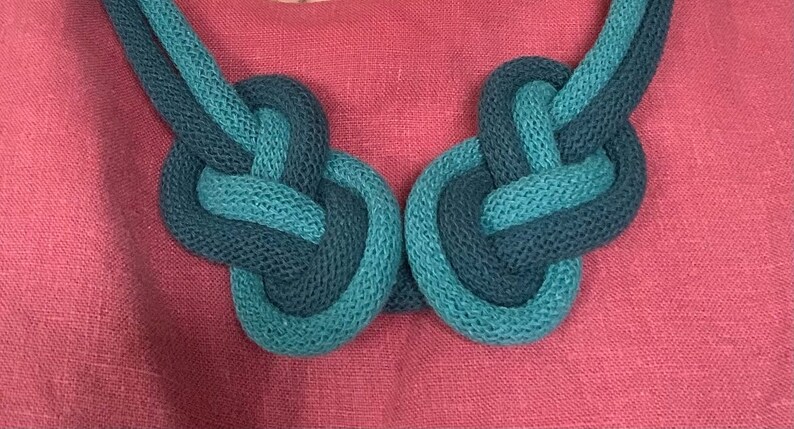 May include: A close-up of a knotted necklace in shades of teal and navy blue. The necklace is made of a thick, textured material and rests against a pink background. The necklace is a statement piece.