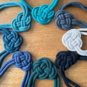 May include: Eight different colored rope knots in the shape of a heart. The knots are made of thick rope and are arranged in a circle. The colors of the knots are teal, dark blue, light blue, black, white, and green.