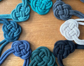 Op de afbeelding: Acht verschillende gekleurde touwknoppen in de vorm van een hart. De knopen zijn gemaakt van dik touw en zijn in een cirkel gerangschikt. De kleuren van de knopen zijn teal, donkerblauw, lichtblauw, zwart, wit en groen.