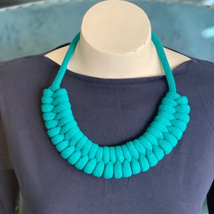 Op de afbeelding: Een turquoise gebreide ketting, tentoongesteld op een mannequin. De ketting is gemaakt van dik, geweven materiaal en vormt een chunky, afgerond ontwerp. De ketting heeft een levendige turkoois tint, en de achtergrond is donkerblauw.