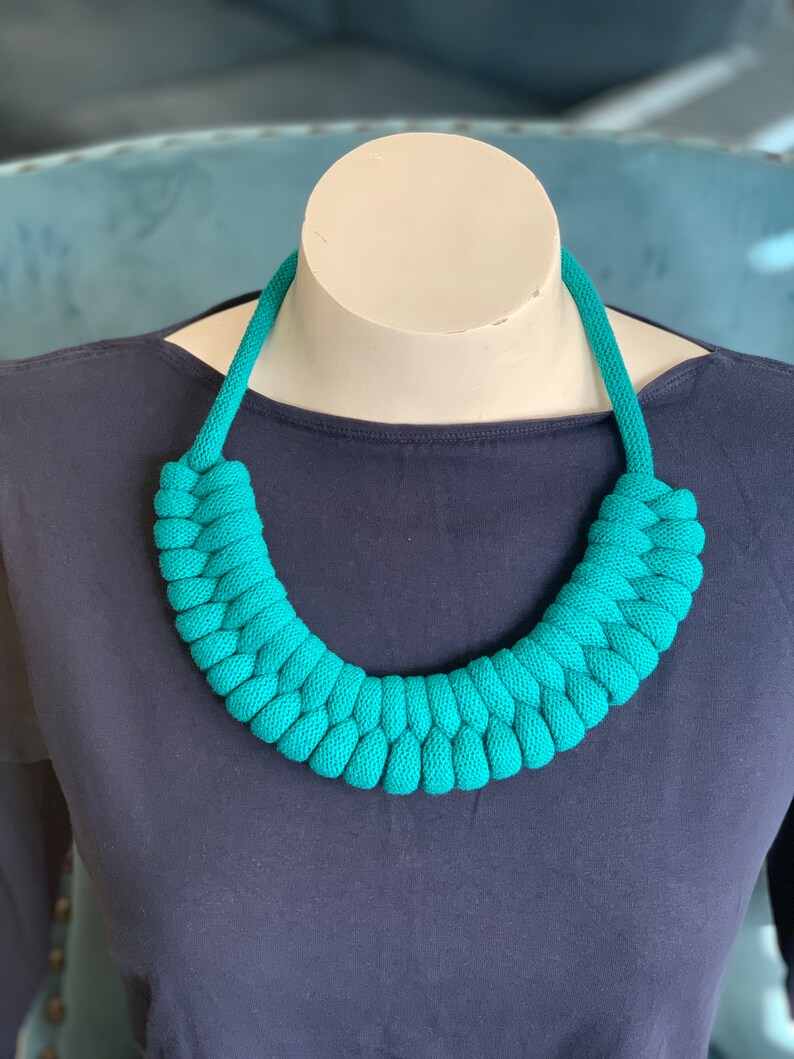 Op de afbeelding: Een turquoise gebreide ketting, tentoongesteld op een mannequin. De ketting is gemaakt van dik, geweven materiaal en vormt een chunky, afgerond ontwerp. De ketting heeft een levendige turkoois tint, en de achtergrond is donkerblauw.