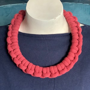 May include: A thick, red, crocheted necklace made of rope. The necklace is displayed on a mannequin wearing a dark blue shirt.