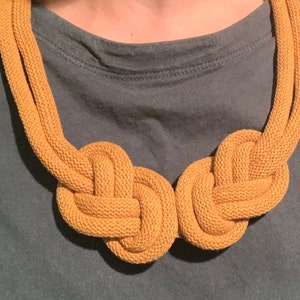 May include: Mustard yellow rope necklace with two large knotted sections. The necklace is made of a thick, textured rope material and is displayed against a teal-colored background. The necklace is a statement piece.