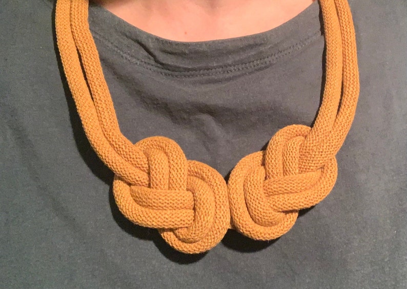 May include: Mustard yellow rope necklace with two large knotted sections. The necklace is made of a thick, textured rope material and is displayed against a teal-colored background. The necklace is a statement piece.