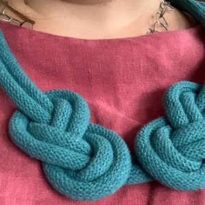 May include: A teal knitted necklace with two large knotted sections. The necklace is made of a soft, textured material and is displayed against a pink background. The necklace is a statement piece.