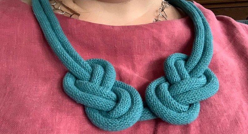 May include: A teal knitted necklace with two large knotted sections. The necklace is made of a soft, textured material and is displayed against a pink background. The necklace is a statement piece.
