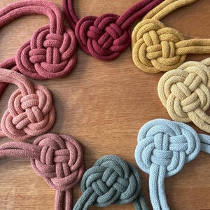 May include: Eight handmade macrame knots in various colors, including red, orange, yellow, green, and blue. The knots are arranged in a circle on a wooden surface.