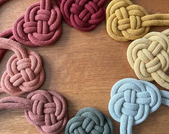 Op de afbeelding: Acht handgemaakte macramé-knopen in verschillende kleuren, waaronder rood, oranje, geel, groen en blauw. De knopen zijn in een cirkel op een houten oppervlak gerangschikt.