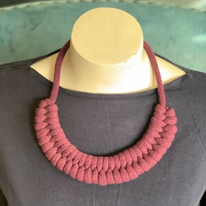 Op de afbeelding: Een bordeauxkleurige ketting met een gevlochten ontwerp. De ketting wordt getoond op een mannequin en wordt gedragen over een donkergrijs shirt met lange mouwen. Het gevlochten gedeelte van de ketting is ongeveer 30 cm breed.