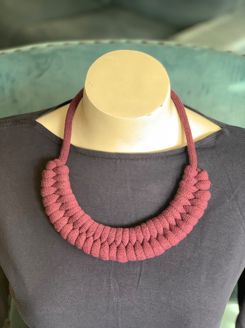 Op de afbeelding: Een bordeauxkleurige ketting met een gevlochten ontwerp. De ketting wordt getoond op een mannequin en wordt gedragen over een donkergrijs shirt met lange mouwen. Het gevlochten gedeelte van de ketting is ongeveer 30 cm breed.