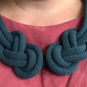 May include: A teal-colored knitted necklace with two large, knotted focal points. The necklace is made of a thick, textured yarn and is displayed against a pink background.