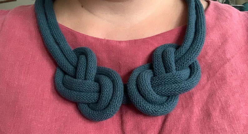 May include: A teal-colored knitted necklace with two large, knotted focal points. The necklace is made of a thick, textured yarn and is displayed against a pink background.