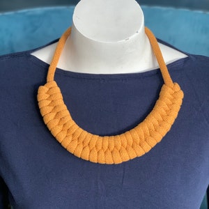 Op de afbeelding: Mosterdgele gebreide ketting op een witte mannequin. De ketting heeft een dik, gevlochten ontwerp en is een opvallend sieraad. De achtergrond is een donkerblauwe top.