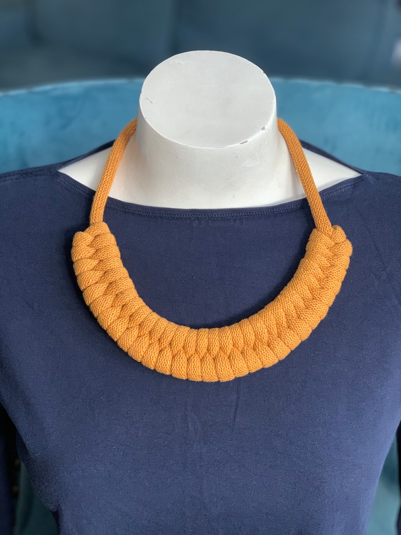 Op de afbeelding: Mosterdgele gebreide ketting op een witte mannequin. De ketting heeft een dik, gevlochten ontwerp en is een opvallend sieraad. De achtergrond is een donkerblauwe top.