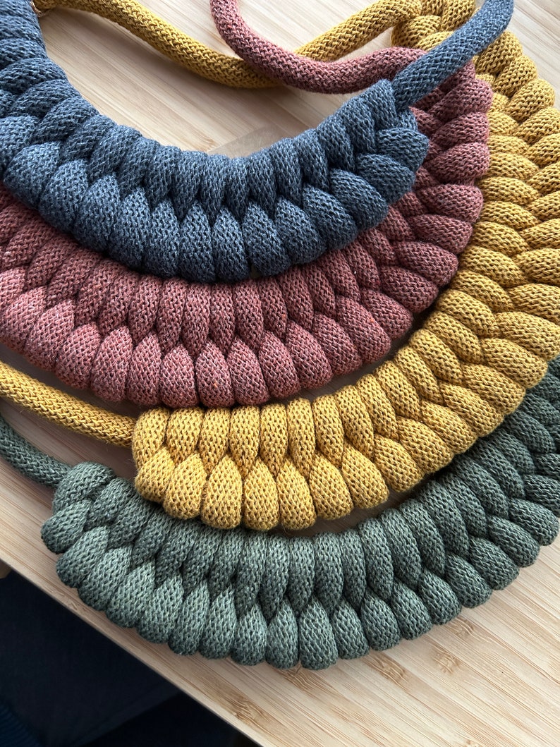 Op de afbeelding: Close-up van verschillende gevlochten touwkettingen in verschillende kleuren, waaronder grijs, stoffig roze, mosterdgeel en olijfgroen. De kettingen zijn gerangschikt in een gelaagd, halfrond patroon, dat hun textuur en ontwerp laat zien. De achtergrond is een lichtgekleurd houten oppervlak.