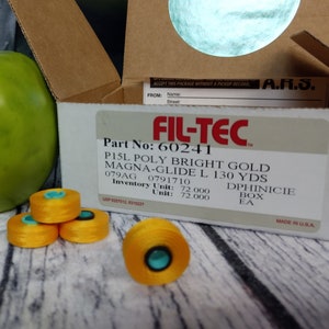 Może przedstawiać: Pudełko z nicią Fil-Tec P15L Poly Bright Gold Magna-Glide L 130 Yds, zielonym jabłkiem i czterema szpulkami żółtej nici na drewnianej powierzchni. Na pudełku widnieje tekst "Part No: 60241" i "Inventory Unit: 72,000".
