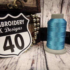 Op de afbeelding: Een spoel lichtblauw borduurgaren met de tekst "Embroidery & Designs 40" op een wit en zwart bord.