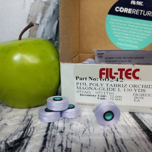 May include: A box of Fil-Tec P15L Poly Tabriz Orchid Magna-Glide L thread in a light purple color. The box has a label with the product information, including the part number 60242, the color code 072AG, and the length of 130 yards. The box also has a label that says "Core Return".