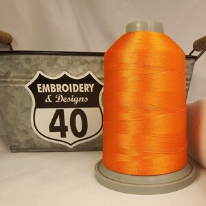 Op de afbeelding: Een spoel met feloranje borduurgaren, met een label met de tekst "Embroidery & Designs 40", die in een metalen container met handvatten ligt.