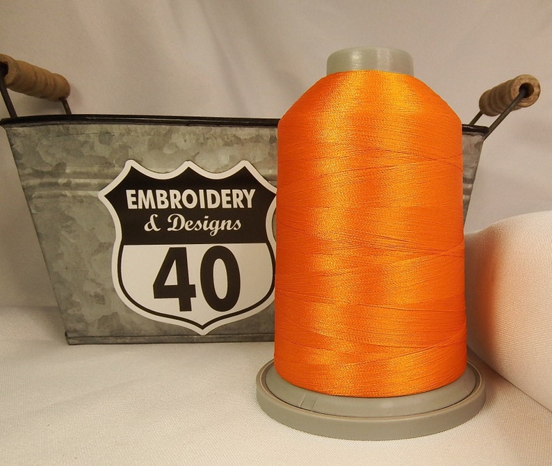 Op de afbeelding: Een spoel met feloranje borduurgaren, met een label met de tekst "Embroidery & Designs 40", die in een metalen container met handvatten ligt.