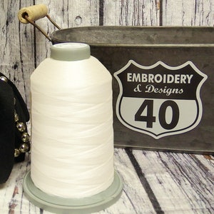 May include: A white spool of embroidery thread with a gray plastic bobbin. The spool is sitting on a gray plastic base. The background is a rustic wooden surface with a metal container with a sign that reads "Embroidery & Designs 40".