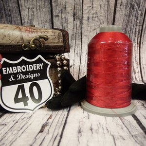 Op de afbeelding: Een spoel rood borduurgaren met een wit label waarop "Embroidery & Designs 40" staat.
