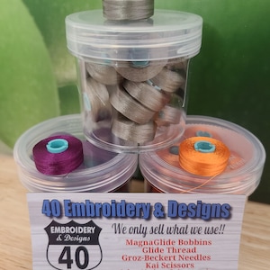 May include: A close-up of a business card for 40 Embroidery & Designs. The card features a red, white, and blue highway sign with the number 40 on it. The text on the card reads "We only sell what we use!!" and lists the products they sell, including MagnaGlide Bobbins, Glide Thread, Groz-Beckert Needles, Kai Scissors, and Select Notions & Accessories. The card also includes contact information, including their website and phone number.