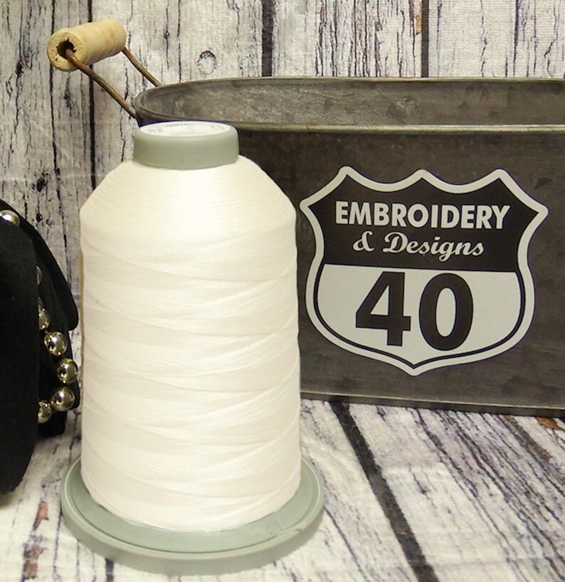 Op de afbeelding: Een witte spoel met borduurgaren op een grijze kunststof spoel. De spoel ligt op een houten oppervlak naast een metalen container met een bord met de tekst "Embroidery & Designs 40".