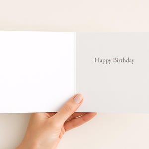 A hand holding an open white greeting card with the text "Happy Birthday" written on it.