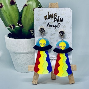May include: A pair of colorful alien-themed earrings with a rainbow-striped design and a black alien head inside a clear UFO. The earrings are on a white card with the text "King Pin Badges" and "@kingpinbadges".