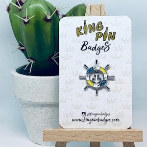 May include: A silver enamel pin with a blue anchor and yellow and white striped wheel. The pin says "We're Anchored" around the wheel. The pin is on a white card with a crown pattern and the text "King Pin Badges" and "www.kingpinbadges.com".