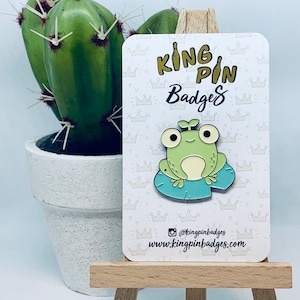 Pode incluir: Um broche de esmalte de sapo verde em um cartão branco com um padrão de coroa. O cartão diz "King Pin Badges" e inclui o endereço do site "www.kingpinbadges.com".