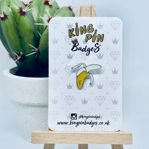 May include: A yellow and white enamel pin in the shape of a peeled banana. The pin is displayed on a card with the text "King Pin Badges" and social media information. A cactus in a white pot is in the background.