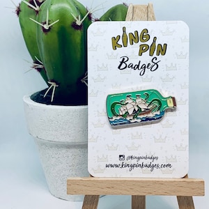 May include: A green enamel pin shaped like a bottle, featuring a ship battling a sea monster. The pin is on a card with the text "King Pin Badges" and social media information. A cactus in a white pot is in the background.