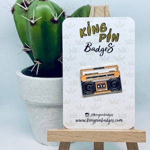 Peut inclure: Une épingle en émail jaune et noire représentant un magnétophone vintage. L'épingle est sur une carte blanche avec un motif de couronne et le texte "King Pin Badges" et "www.kingpinbadges.com".