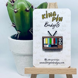 May include: A retro television enamel pin with a color bar test pattern. The pin is brown and black with a silver antenna and two speaker holes. The pin is on a white card with the text "KING PIN Badges".