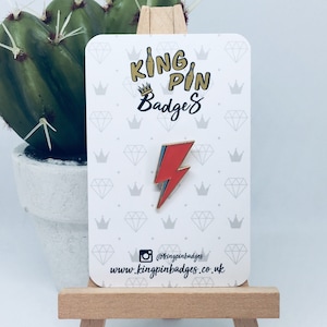 Peut inclure: Épingle en forme d'éclair rouge et bleu avec bordure dorée, présentée sur une carte blanche avec le texte "King Pin Badges". La carte est posée sur un petit chevalet en bois, avec un cactus en pot blanc en arrière-plan.
