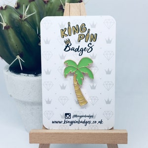 May include: A palm tree enamel pin with a gold outline and a light green frond. The pin is on a white card with the text "KING PIN Badges" and social media information. The pin is displayed on a small wooden easel.